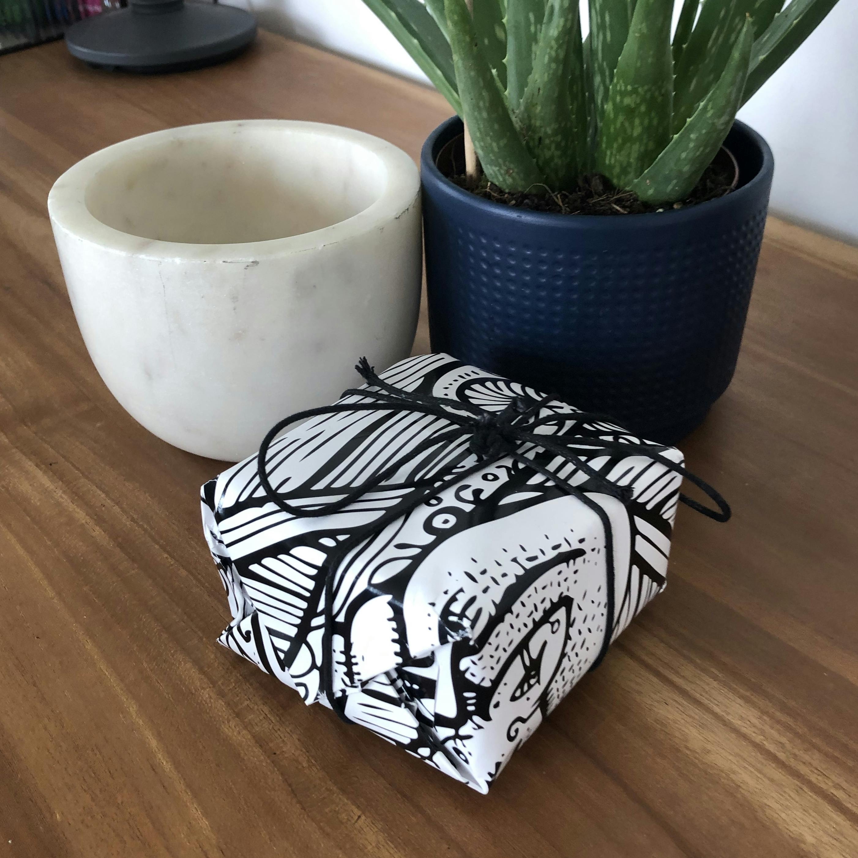 Photograph of a little box nicely wrapped up in glossy, black and white paper. There's a plant and a heavy kitchen mortar in the composition too, all on top of a wooden desk